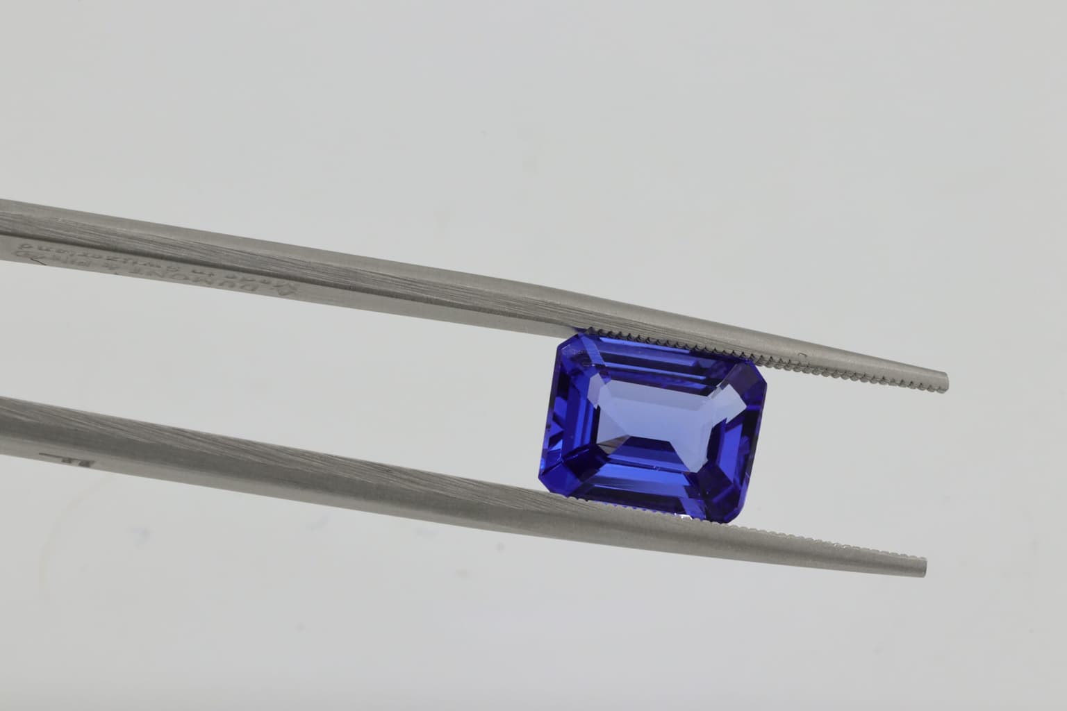 A violet-blue emerald-cut gemstone held in a pair of gemmologist's tweezers against a plain pale background